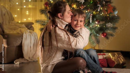 Positive smiling mother and son hugging and kissing on Christmas eve in living room. Families and children celebrating winter holidays.