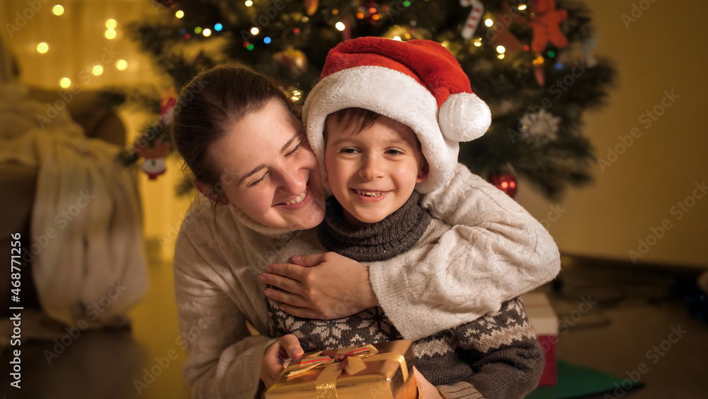 Smiling mother giving Christmas present in golden box to her smiling boy in Santa hat. Families and children celebrating winter holidays. Smiling mother giving Christmas present in golden box to her smiling boy in Santa hat. Families and children celebrating winter holidays.