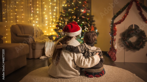 Rear view of hugging mom with son in warm wool sweater sitting next to glowing Christmas tree