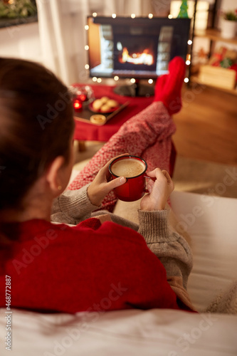 christmas, winter holidays and leisure concept - close up of young woman watching tv with fireplace on screen and drinking coffee with her feet on table at cozy home