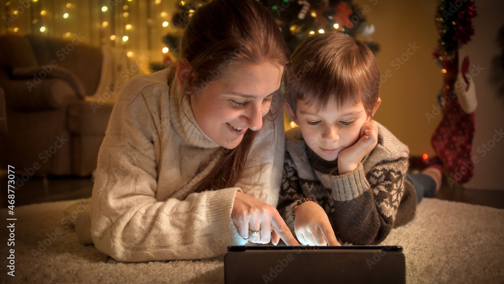 Portrait of smiling mom with son browsing internet and watching video on tablet under Christmas tree. Pure emotions of families and children celebrating winter holidays. Portrait of smiling mom with son browsing internet and watching video on tablet under Christmas tree. Pure emotions of families and children celebrating winter holidays.