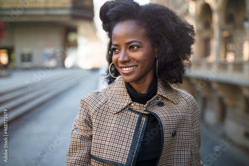 Beautiful African American young woman with afro and large hoop earrings in a stylish coat, smiling. Urban street portrait. Spring or fall time. Selective focus, copy space