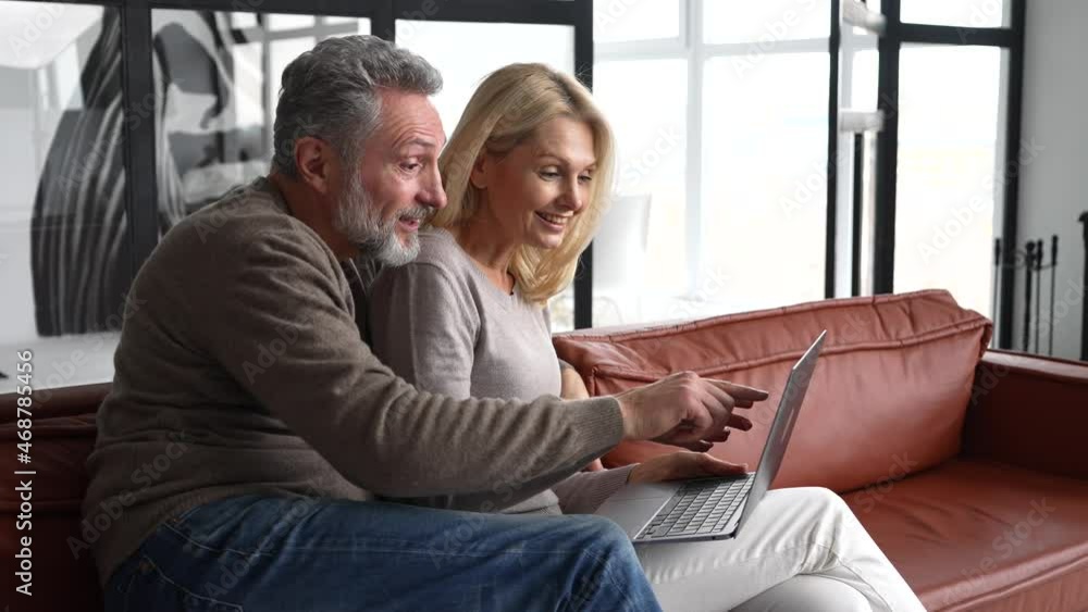 Middle-aged couple spends leisure time with a laptop at home. Spouses sit on the couch, watching video, web surfing or shopping online together, enjoying time online Middle-aged couple spends leisure time with a laptop at home. Spouses sit on the couch, watching video, web surfing or shopping online together, enjoying time online
