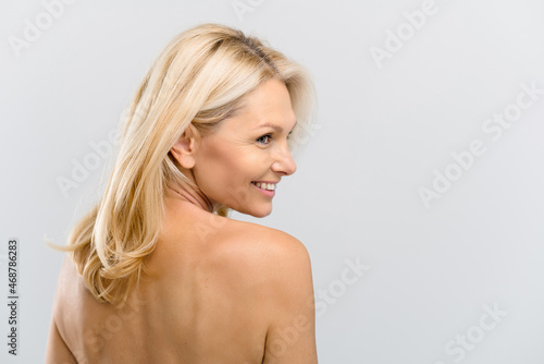 Joyful and happy middle-aged woman posing over grey background. Charming half-naked mature lady with clean pure skin turning around and laughing. Anti-aging treatment and body care concept