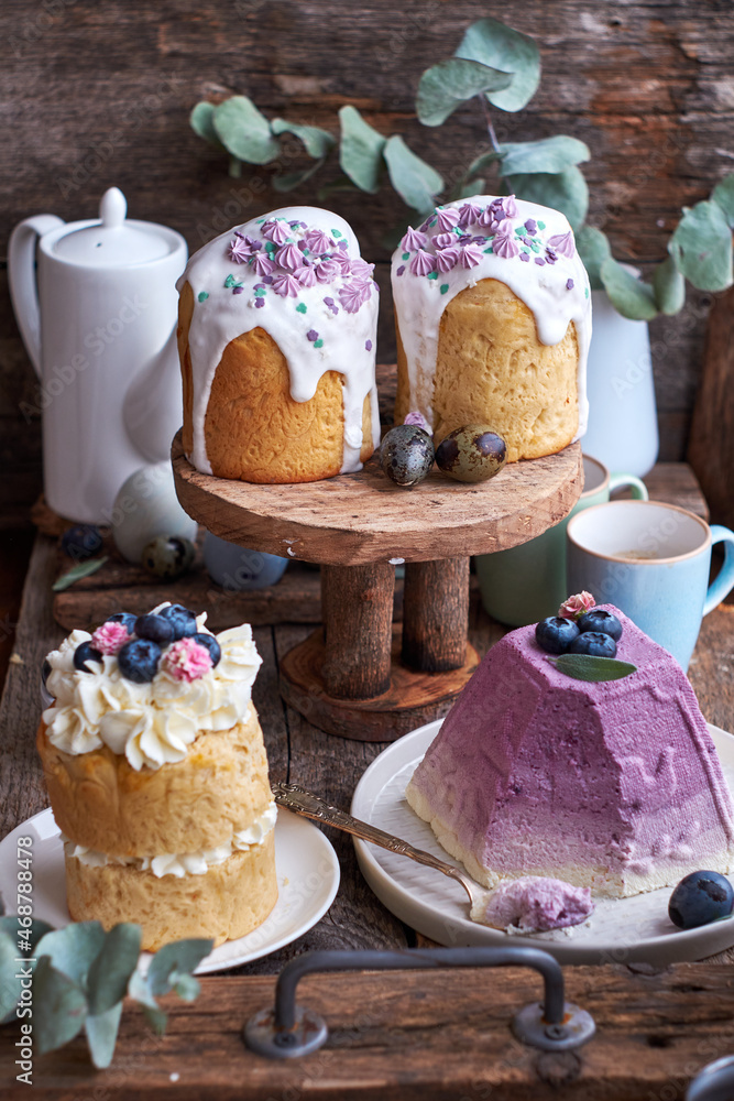 Easter cakes with glaze and decorated mini meringue, cream, blueberries and Traditional Easter Orthodox curd cake with blueberry. Wooden background, side view Easter cakes with glaze and decorated mini meringue, cream, blueberries and Traditional Easter Orthodox curd cake with blueberry. Wooden background, side view