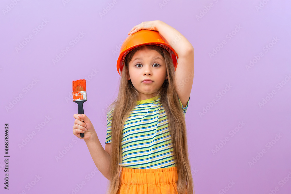 A little girl holds a construction helmet on her head with her hand, and with her other hand holds a paint brush for painting walls in a children's room. A little girl holds a construction helmet on her head with her hand, and with her other hand holds a paint brush for painting walls in a children's room.
