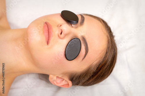 Young woman receiving facial massage with black mineral stones on her eyes in a spa