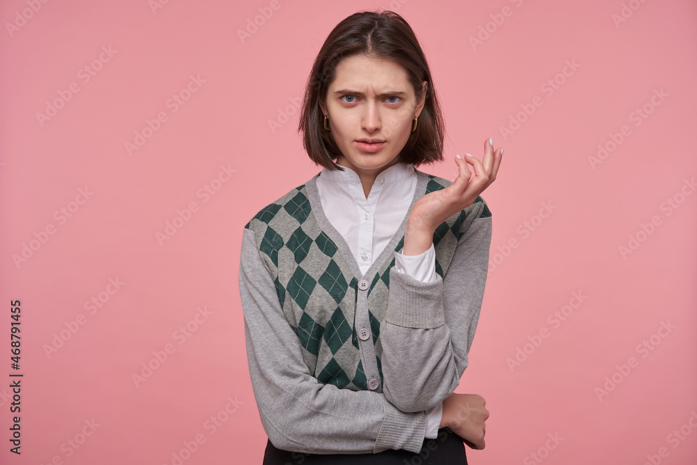 Indoor shot of young brunette student female, starring into camera with angry, mad facial expression, keeps her hand folded. Isolated over pink background. Indoor shot of young brunette student female, starring into camera with angry, mad facial expression, keeps her hand folded. Isolated over pink background.