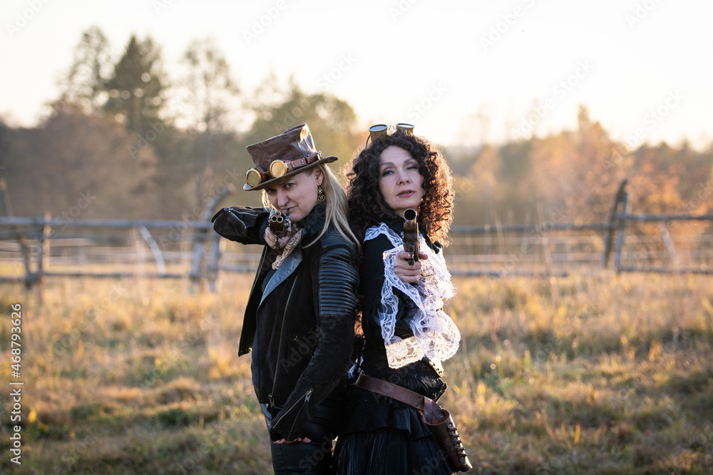 Two women in steampunk suits with fake pistols stand back to back against an autumn landscape at a ranch. They aim directly at the camera