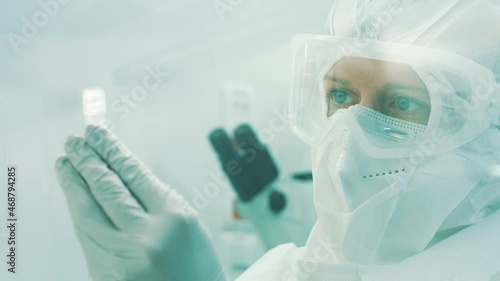 Close up shot of a Caucasian female researcher in a full protective suit, with glasses, face mask, and gloves, holding and looking at a cryogenic tube. photo