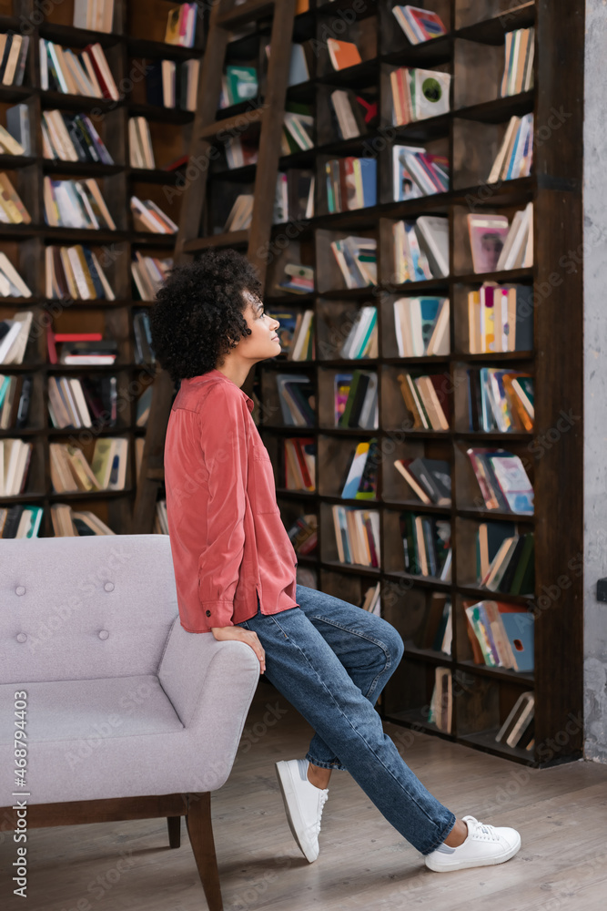 side view of african american woman leaning on grey couch near bookshelf side view of african american woman leaning on grey couch near bookshelf