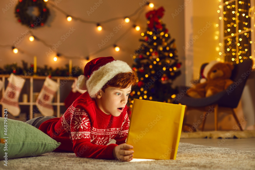 Surprised boy in Santa hat opens magic book, reads Christmas stories and fairy tales, discovers wonderful things, and, like every little child, believes in miracles and hopes his Xmas dream comes true