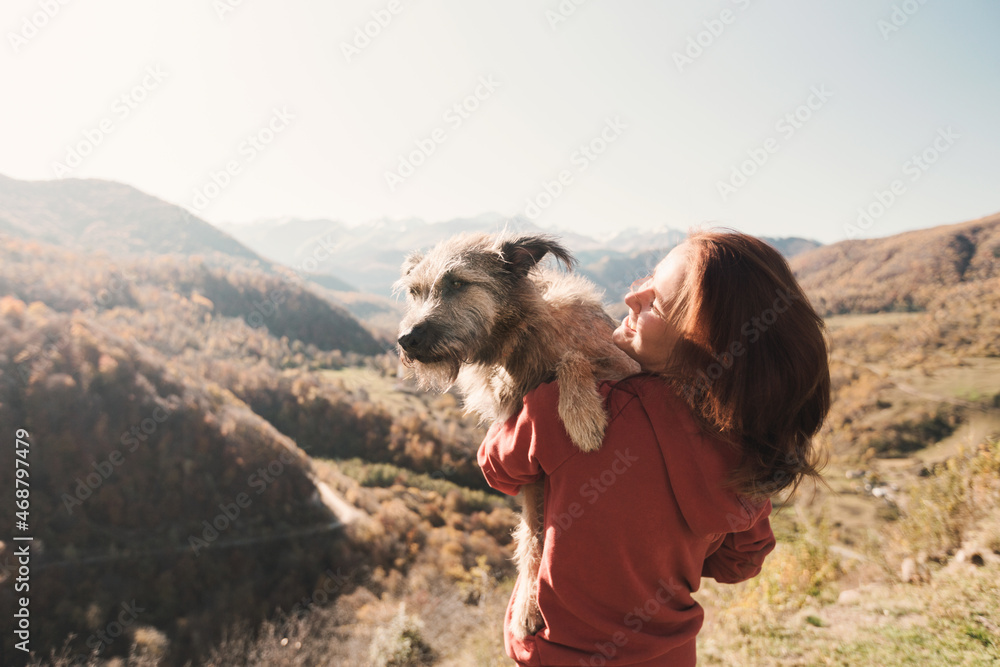 Happy woman and her pet. She is holding a dog in her arms, sunny day, mountains.