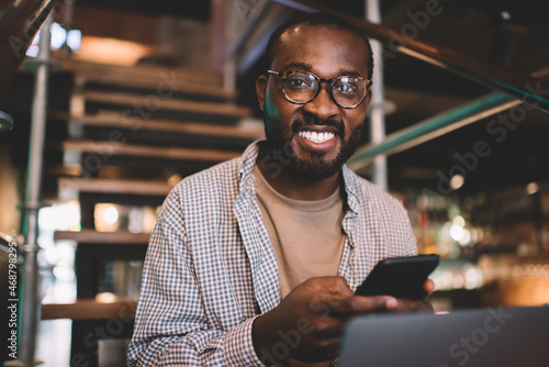 Portrait of cheerful male blogger with cellphone gadget and cropped laptop smiling at camera during free time, happy African American man in classic glasses using smartphone and netbook indoors