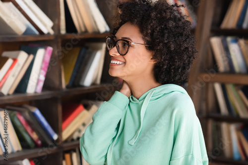 cheerful african american student in eyeglasses looking at bookshelf