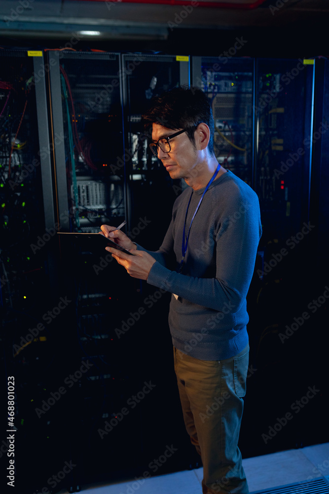 Asian male engineer writing on clipboard in computer server room
