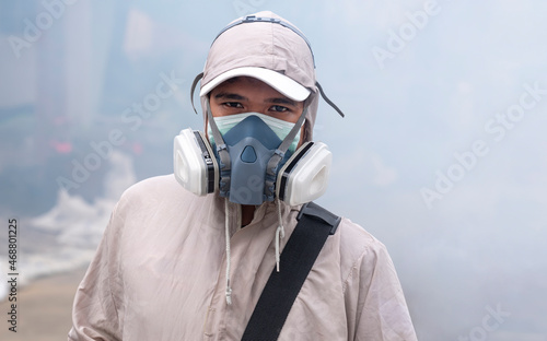 Asian outdoor healthcare worker in multi-purpose respirator half mask with protective clothing looking at camera on chemical fume background