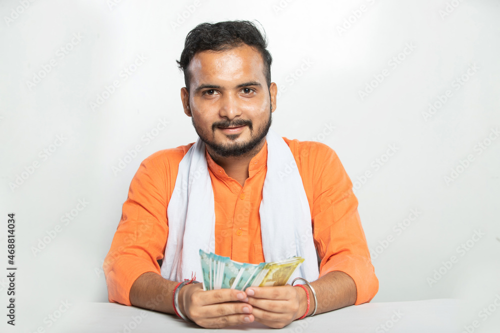 Headshot of a young Indian rural mustache man wearing orange kurta sitting against isolated white background holding money in hands, happy village male with beard smiling counting cash notes. Headshot of a young Indian rural mustache man wearing orange kurta sitting against isolated white background holding money in hands, happy village male with beard smiling counting cash notes.