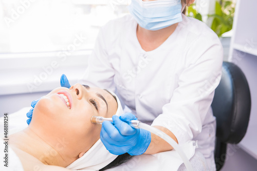 Cosmetologist in a protective medical mask and gloves making the facial microdermabrasion rejuvenation procedures to her client in a beauty salon.