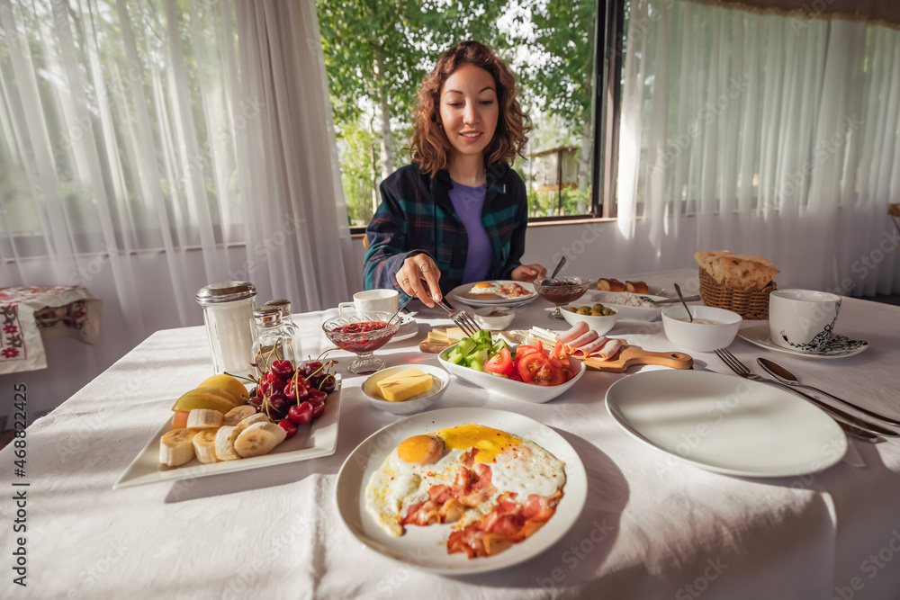 A woman eats a hearty and plentiful continental breakfast in a hotel or cafe in the morning. A plate of fried eggs in the foreground
