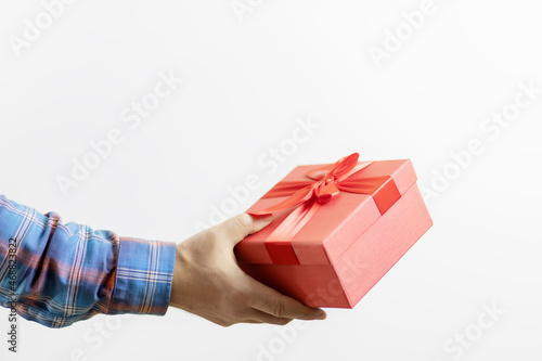 A man's hand in a checkered sleeve holds a red box with a gift on a white background. Gift for a celebration
