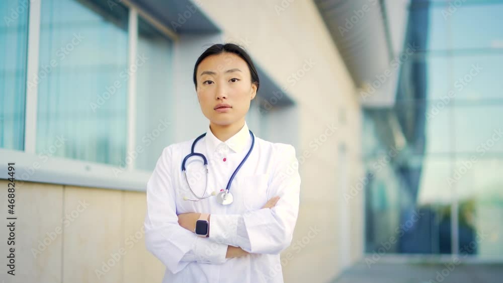 Close up portrait of Asian woman doctor looking at camera with arms crossed on background of modern hospital outside. Confident medic outdoor. female scientist therapist wearing scrubs near clinic Close up portrait of Asian woman doctor looking at camera with arms crossed on background of modern hospital outside. Confident medic outdoor. female scientist therapist wearing scrubs near clinic