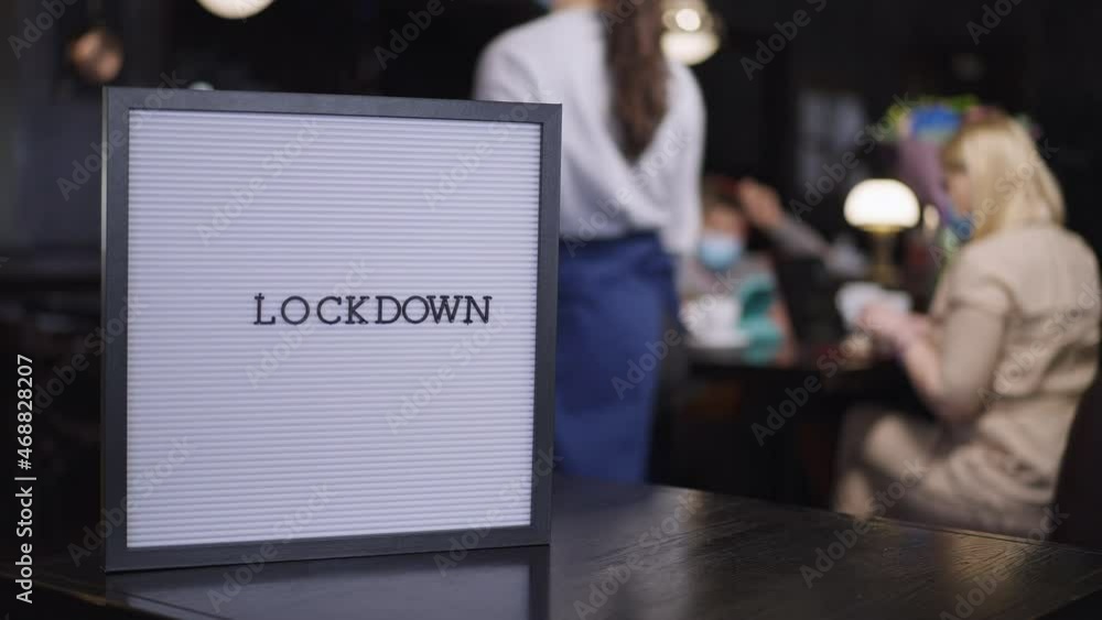 Close-up Lockdown announcement in restaurant with blurred family in face masks dining at background and waitress passing in slow motion. New normal service in cafe indoors on coronavirus pandemic Close-up Lockdown announcement in restaurant with blurred family in face masks dining at background and waitress passing in slow motion. New normal service in cafe indoors on coronavirus pandemic