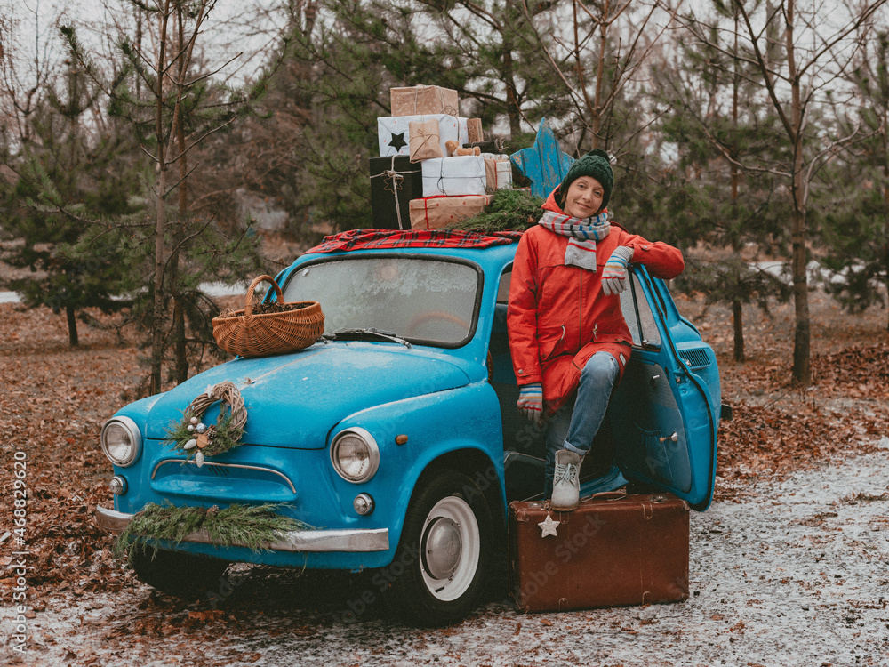 young cheerful female holding Christmas gifts against background of a blue retro car on New Year's Eve