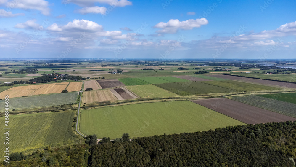 Luftaufnahme von flachem Land mit vielen Feldern und Wald bei blauem Himmel und Windrädern im Hintergrund