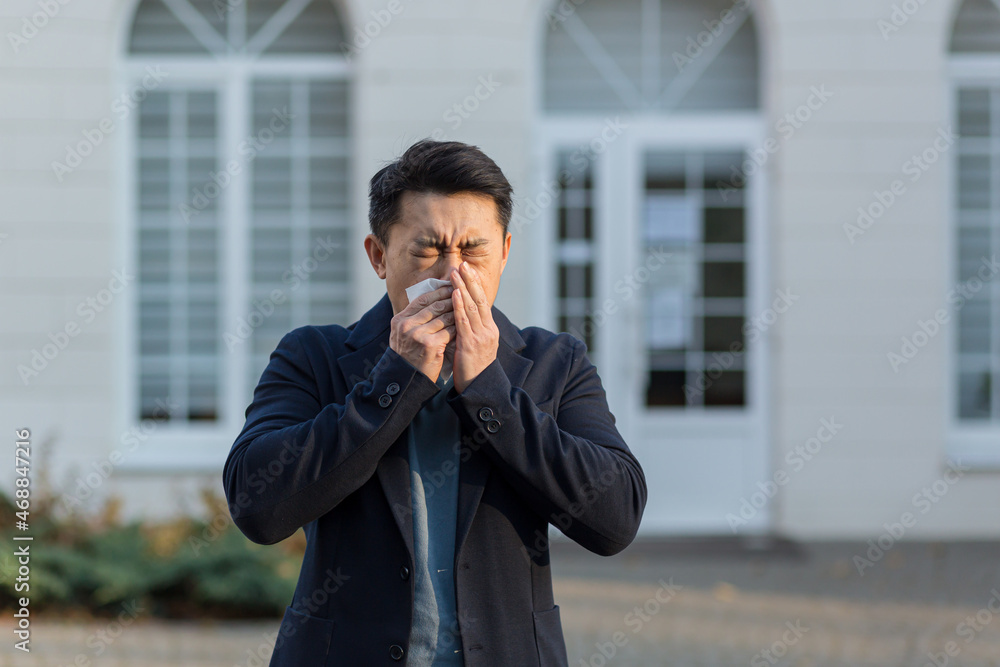 An Asian sick man sneezes near the office on the street, has allergies and illness An Asian sick man sneezes near the office on the street, has allergies and illness