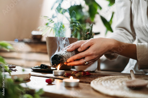 Woman hands burning white sage, palo santo before ritual on the table with candles and green plants. Smoke of smudging treats pain and stress, clear negative energy