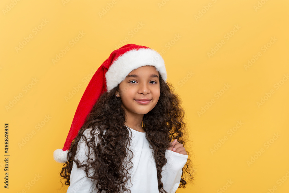 Studio shor of dark skinned young girl with long curly hair in a Santa Claus hat on a yellow background Studio shor of dark skinned young girl with long curly hair in a Santa Claus hat on a yellow background
