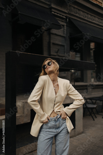 Young beautiful blonde posing effectively with her head thrown back, holding her hands in jeans pockets. Girl in black glasses and beige jacket stands against backdrop of dark building of street.