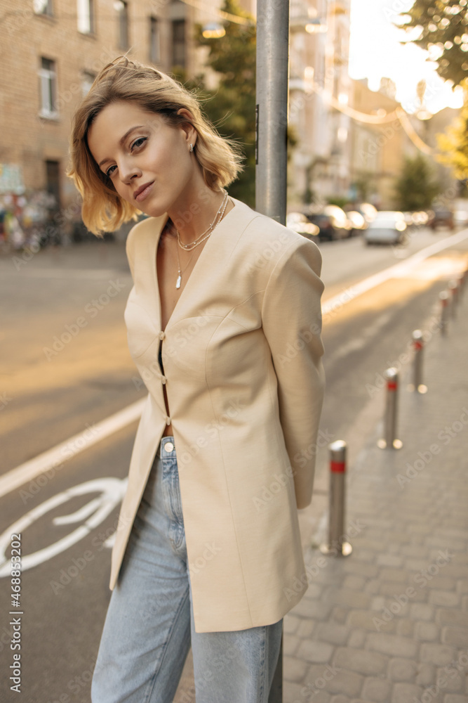 Stylish image of attractive young woman standing on city street holding on to pillar from behind. Light-skinned lady with bob haircut in jeans and beige coat buttoned up.