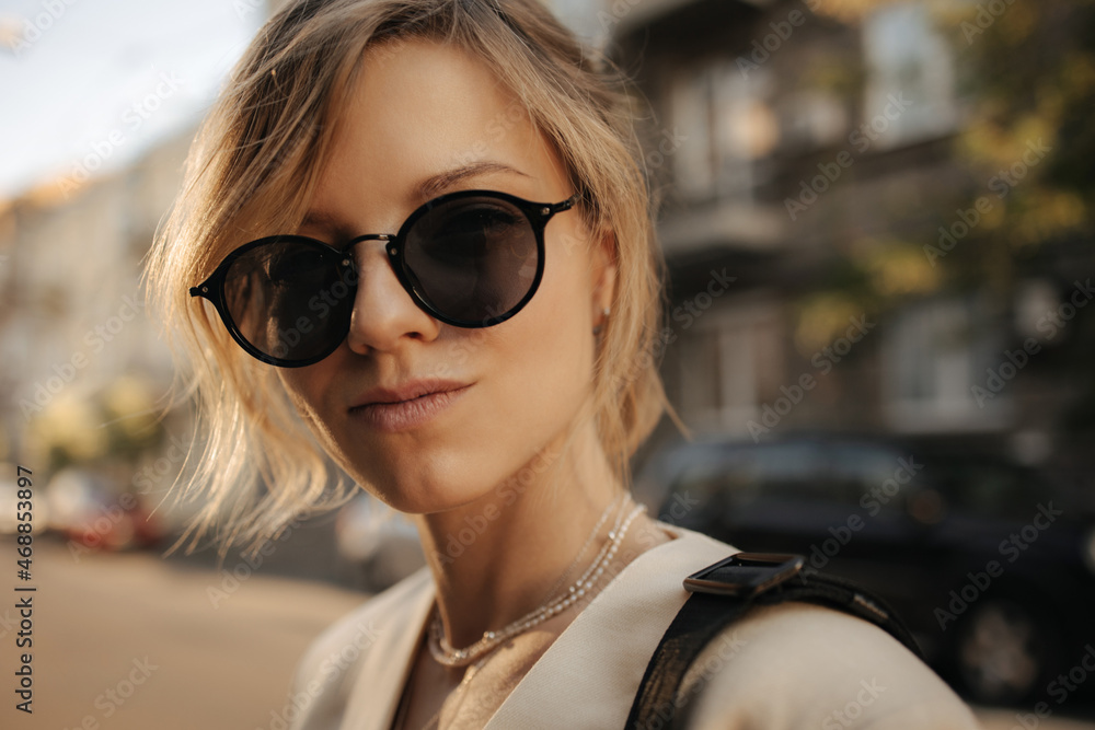 Close up portrait of caucasian woman in black sunglasses on blurred background. Blonde with gathered hair narrows her eyes and purses her lips, she is wearing white necklace and beige jacket.