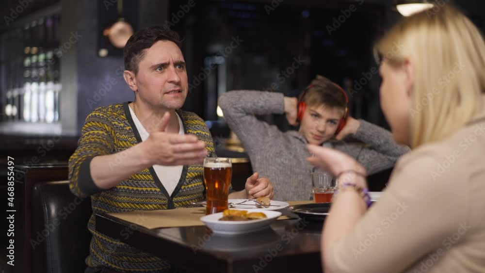 Angry couple of parents arguing in restaurant with blurred teenage son in headphones sitting at background. Portrait of unhappy Caucasian family yelling gesturing shouting in slow motion