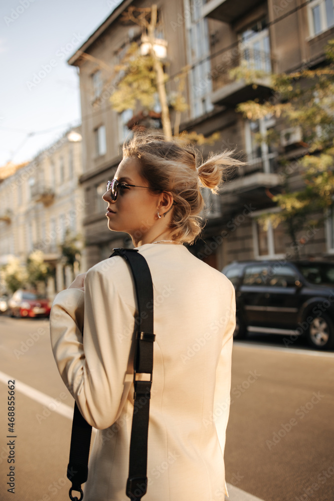 Fototapeta premium Rear view of stylish fair-haired girl in black glasses walking along city street looking to side. Her short hair is pulled back in bun, wearing white cashmere jacket and shoulder bag.
