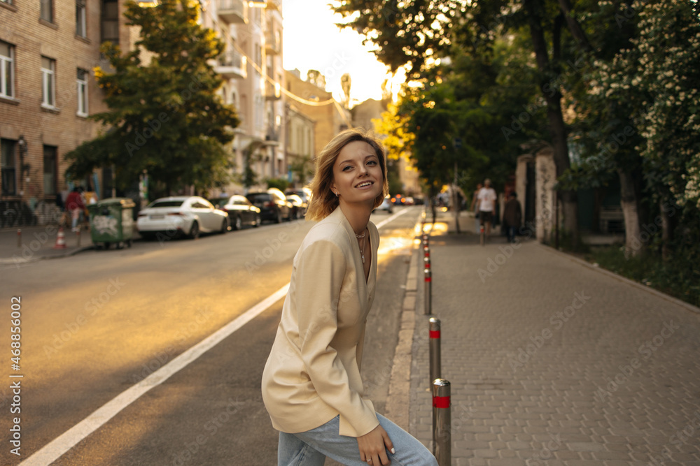 Fototapeta premium Not full-length caucasian young woman cute smiling on street. Girl with short hair crosses road, looking around in beige jacket and light-colored jeans. Warm day for walk in fresh air.