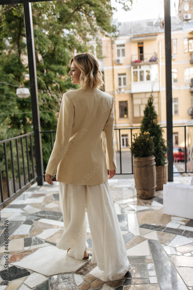 Full-length tall happy caucasian young woman stands with her back to camera on terrace during daytime. Blonde with short wavy hair in wide white pants and long beige jacket.