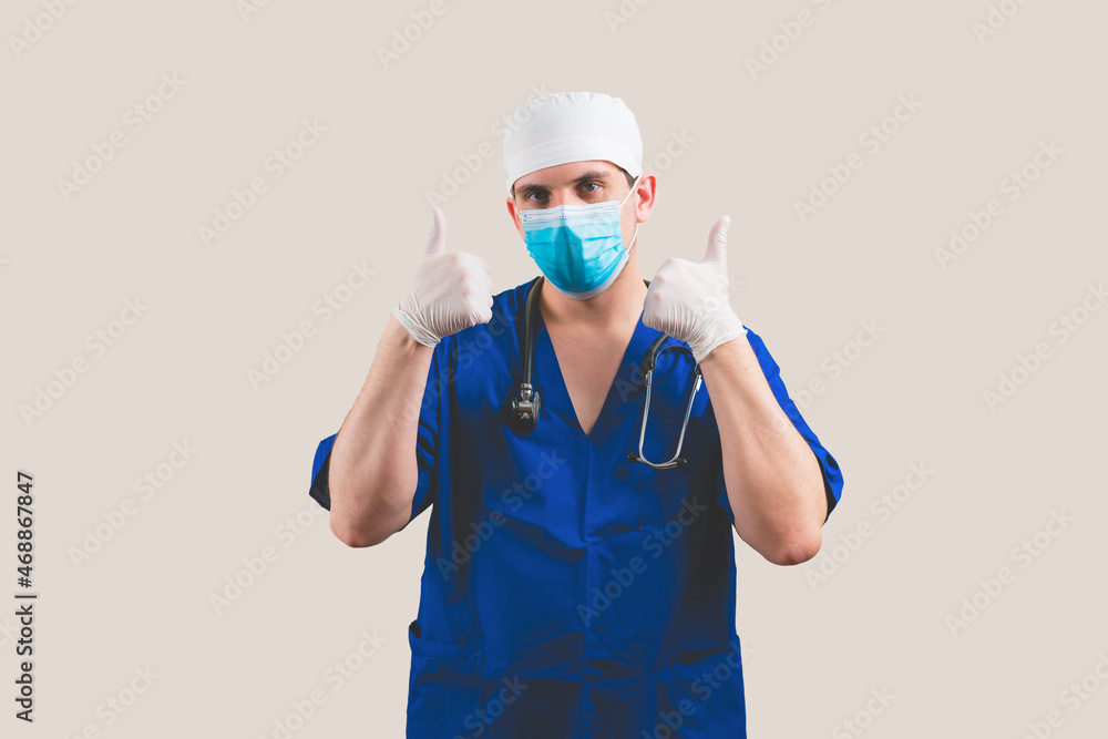 Young doctor with thumbs up in blue surgeon suit and glasses with stethoscope on the yellow background isolated Young doctor with thumbs up in blue surgeon suit and glasses with stethoscope on the yellow background isolated