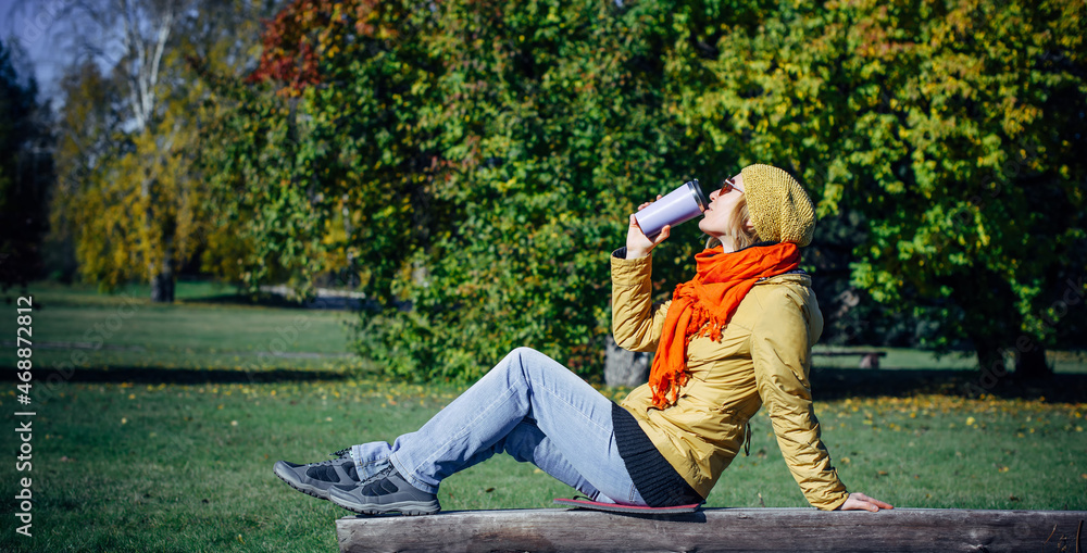 Naklejka premium Pretty european woman in yellow jacket and red scarf sitting on wooden bench in a deserted park and drinking coffee. Enjoying solitude and sunny autumn weather. Blurred plant background, copy space.
