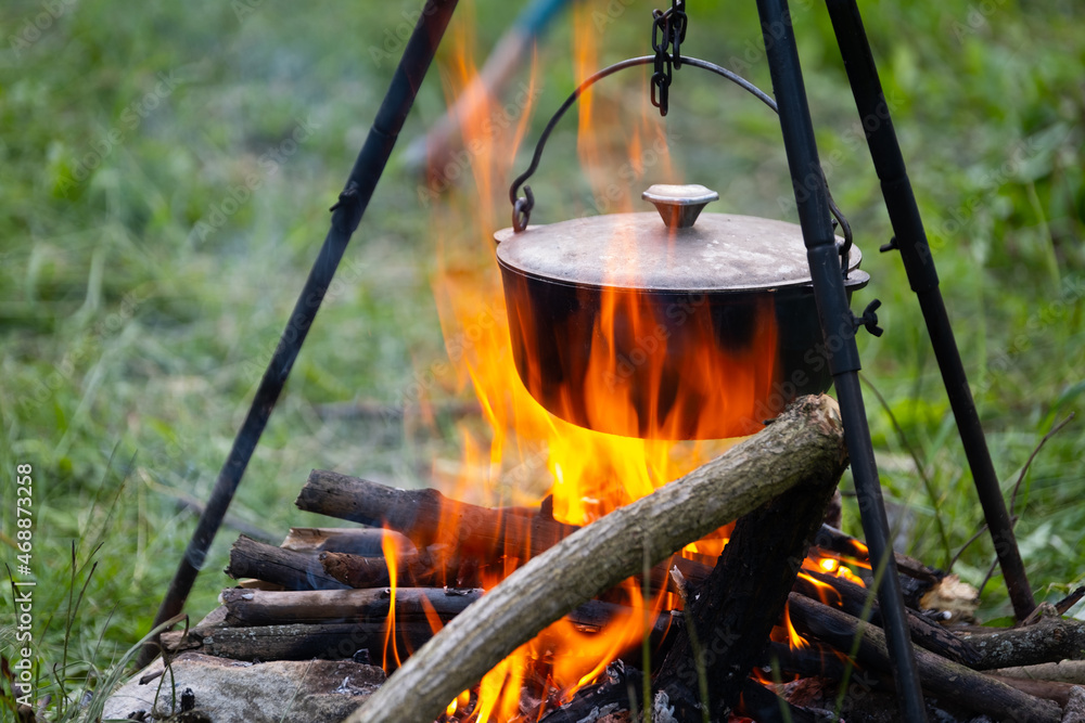 Black large tourist pot hangs over burning flame of campfire in the forest.  Cooking dinner on