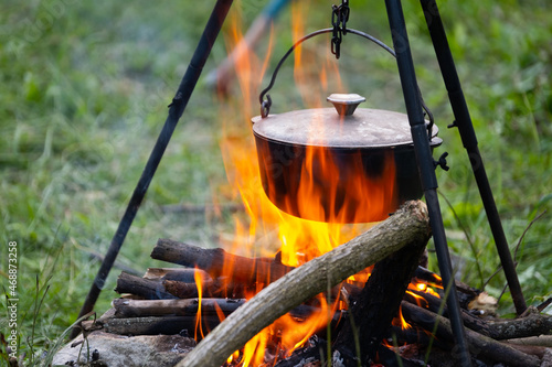 Black large tourist pot hangs over burning flame of campfire in the forest. Cooking dinner on wild hike outdoor. Cauldron of hot food is heated over fire. Outdoor travel. Boil of hot meal in camp.
