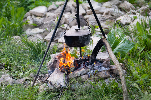 Black large tourist pot hangs over burning flame of campfire in the forest. Cooking dinner on wild hike outdoor. Cauldron of hot food is heated over fire. Outdoor travel. Boil of hot meal in camp.