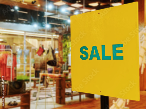Sale Promotional Text on Yellow Label Against Blurred Clothing Store Background