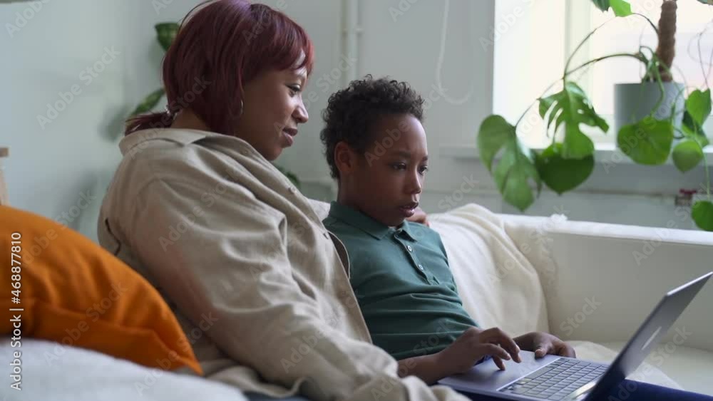 Distant home schooling of junior children. Little African-American boy pupil uses laptop to spbd study online with mother on couch against window with pot plant Distant home schooling of junior children. Little African-American boy pupil uses laptop to spbd study online with mother on couch against window with pot plant