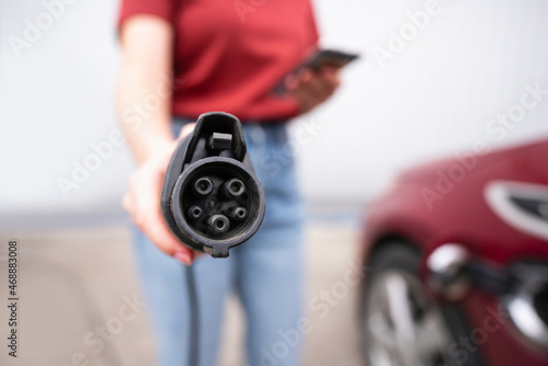 Woman with smartphone shows electric car charging plug at camera