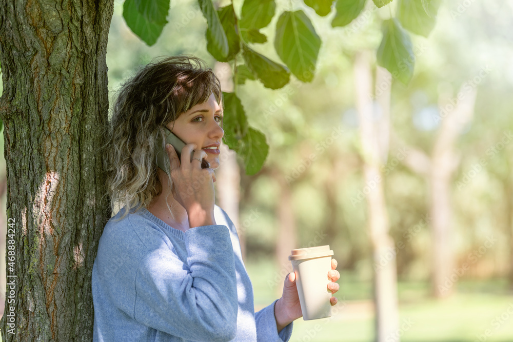 Woman leaning in a tree and talking with mobile with a take away coffee in hand