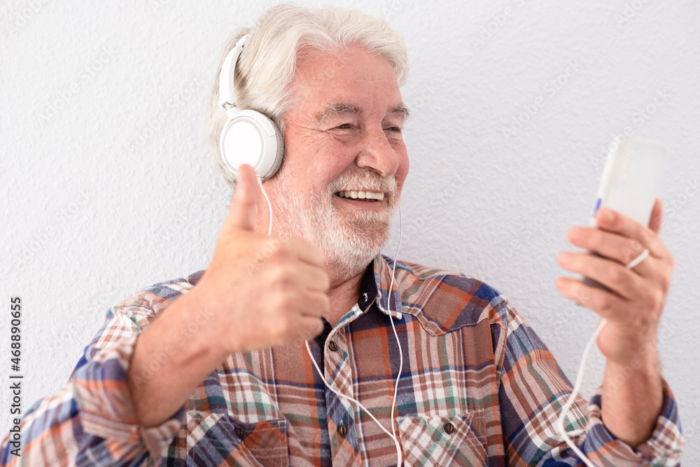 Smiling senior man with white hair using mobile phone in video chat wearing headphones. Gesturing a positive sign with hand Smiling senior man with white hair using mobile phone in video chat wearing headphones. Gesturing a positive sign with hand