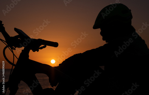 Silhouette of man with bicycle sitting on the cliff wearing helmet after cycling along the sea. Orange-black background